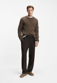 Maglione marrone a maglia con scollo a girocollo indossato sopra una camicia bianca, abbinato a pantaloni plettati marrone scuro e sneakers beige.