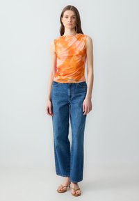 Oranje tie-dye mouwloze top met gathered detail, gecombineerd met high-waisted blauwe jeans en tan sandalen, staande tegen een neutrale achtergrond.
