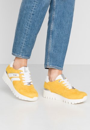 Baskets en daim jaune avec semelles et lacets blancs portées avec un jean bleu raccourci sur un sol gris clair.