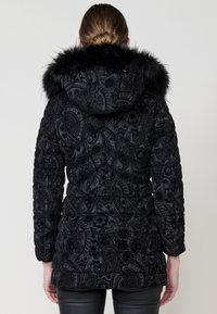 Svart quiltad puffjacka med paisleymönster, fuskpälsfodrad huva och långa ärmar, med en slät och texturerad yta.