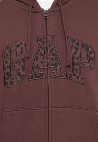 Maroon zip-up hoodie featuring a large "GAP" logo in animal print. Soft texture with drawstring hood and front pocket.