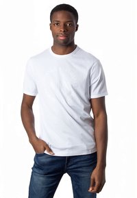 Άνδρας που φοράει λευκό γραφικό μπλουζάκι και μπλε τζιν, στέκεται με το ένα χέρι στην τσέπη μπροστά από λευκό μονόχρωμο φόντο.