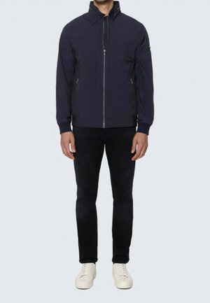 Uomo che indossa una giacca blu navy con zip e collo alto, pantaloni neri e scarpe da ginnastica bianche, in piedi davanti a uno sfondo chiaro e semplice.