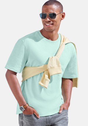 Mann trägt hellblaues T-Shirt und Sonnenbrille, mit einem beigen Sweatshirt um die Schultern gebunden und Händen in den Taschen der grauen Jeans.