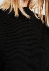 Svart ribbad stickad topp med hög halsringning. Texturen har vertikala linjer och tyget verkar mjukt ochStretch.