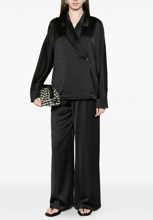 Donna che indossa una giacca doppiopetto larga in raso nero e pantaloni a gamba larga, tenendo una piccola clutch con motivo geometrico bianca e nera.