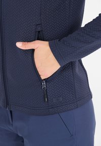 Marinblå jacka med vävt mönster, fickor med dragkedja och en figursydd design. Har en logotypetikett och ett mjukt, stretchigt material.