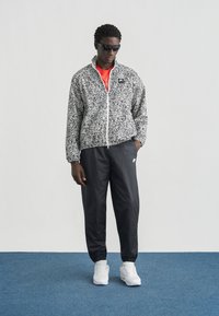 Αδιάβροχο μπουφάν με μαύρο και άσπρο πουά μοτίβο, κλείσιμο με φερμουάρ, συνδυασμένο με μαύρο παντελόνι φόρμας και λευκά αθλητικά παπούτσια, φορεμένο με γυαλιά ηλίου.