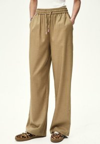 Osoba oblečená v voľných svetlohnedých nohaviciach so šnúrkou v pätách a leopardím vzorom na slip-on topánkach, stojaca na jednoduchom svetlom pozadí.