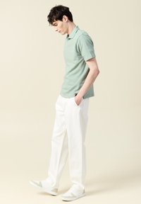 Vihreä teksturoitu poolopaita, valkoiset housut ja valkoiset lenkkarit harmailla yksityiskohdilla. Malli seisoo käsi taskussa ja katsoo alas.