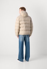 Beige puffer jacket with a hood, featuring horizontal quilted sections. Worn over blue wide-leg jeans with subtle fading, complemented by black shoes.