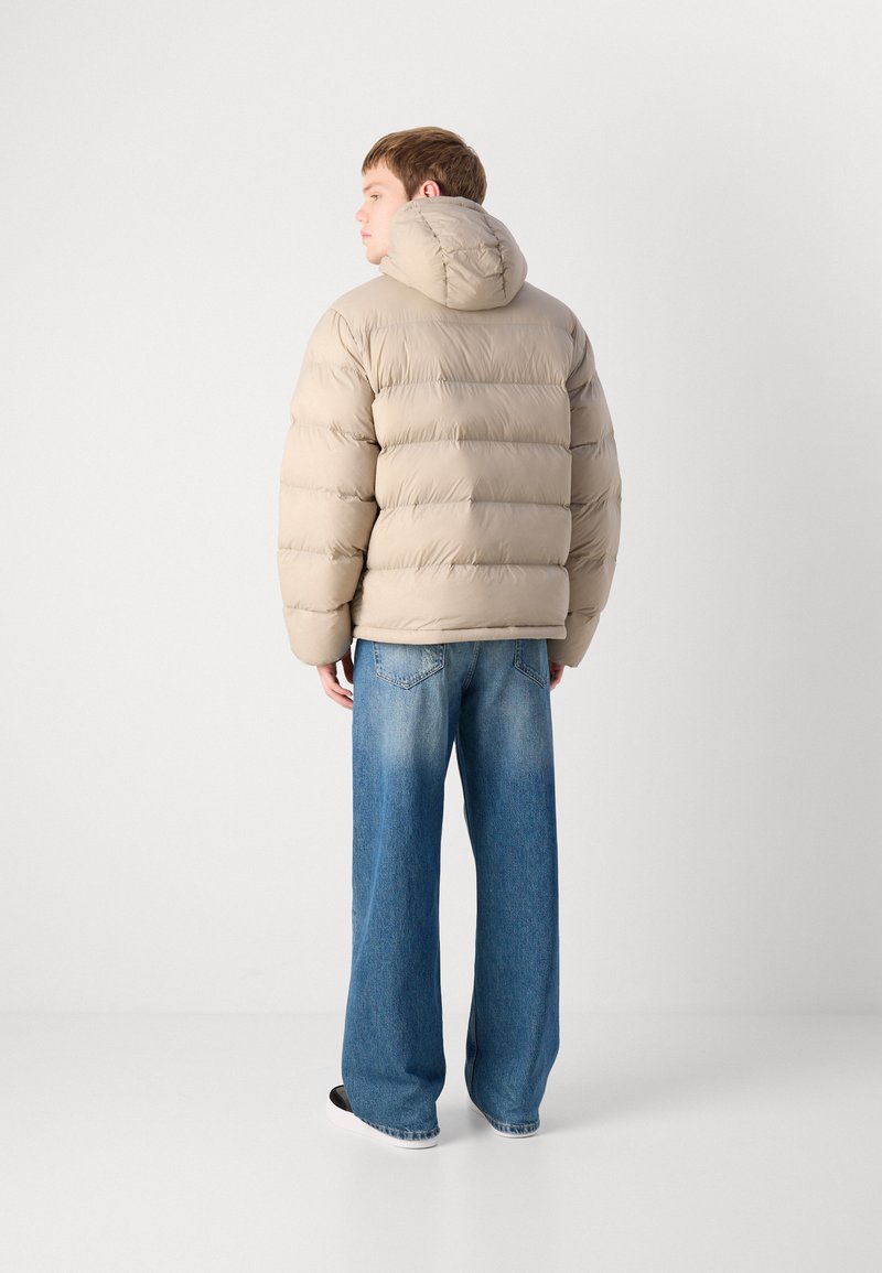 Veste doudoune beige avec capuche, présentant des sections matelassées horizontales. Portée sur un jean large bleu avec un léger délavage, complété par des chaussures noires.