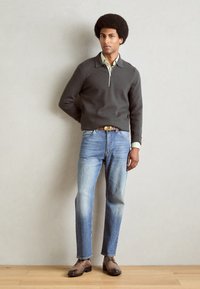 Maglione grigio scuro con zip, camicia verde chiaro sotto, jeans di denim blu e scarpe di pelle marrone, in piedi contro uno sfondo semplice.