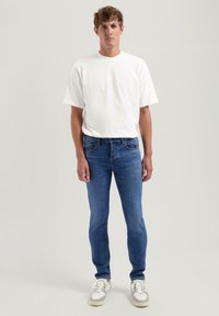Bela kratka majica s kratkimi rokavi v kombinaciji s slim-fit modrimi kavbojkami in belimi športnimi copati. Oblačilo ima preprost, sproščen dizajn s čistimi linijami.