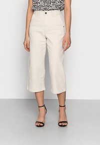 Mujer vestida con pantalones cortos de pierna ancha de cintura alta en color beige y tacones negros con tiras, de pie sobre un suelo gris claro contra una pared blanca.