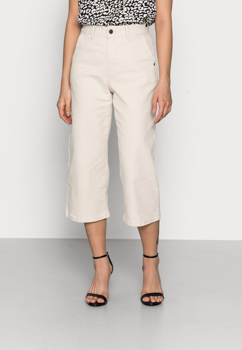 Mujer vestida con pantalones cortos de pierna ancha de cintura alta en color beige y tacones negros con tiras, de pie sobre un suelo gris claro contra una pared blanca.