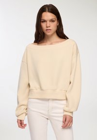 Sudadera cropped color crema con spalle basse, scollatura ampia e orlo a coste. Tessuto morbido, vestibilità leggermente ampia e maniche lunghe.