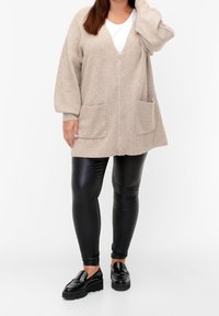 Beżowy dzianinowy kardigan z zamkiem, z dekoltem w serek i dwoma przednimi kieszeniami, zestawiony z czarnymi leginsami i masywnymi czarnymi loafersami, na białym tle.