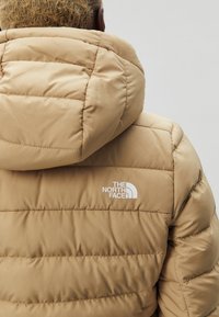 Veste matelassée isolée beige avec capuche, présentant des coutures diagonales et un logo blanc sur l'épaule gauche. Texture lisse.