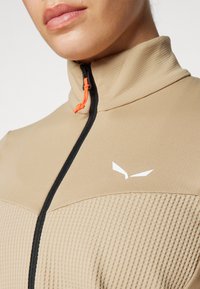 Beige jakke med høy krage, kontrastfylt svart glidelås og oransje snor. Teksturert stoff med en hvit logo på brystet.