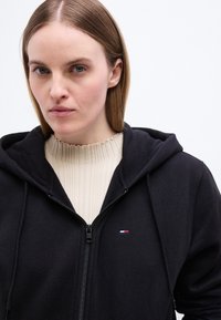 Γυναίκα με ίσια ανοιχτό καστανά μαλλιά φοράει μαύρη ζακέτα με φερμουάρ πάνω από εκρού ριμπ λεπτομέρεια πουλόβερ, κοιτάζοντας μπροστά με ουδέτερη έκφραση.