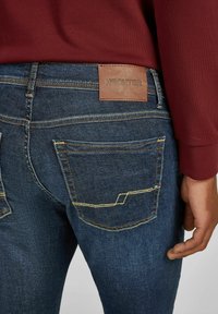 Vue arrière d'une personne portant un jean bleu foncé avec des coutures jaunes et un patch en cuir marron portant la mention "HECHTER".