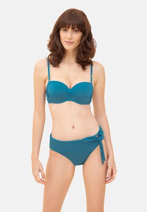 Kvinde med skulderlangt brunt hår iført en to-delt badedragt i teal med bandeau-top og bund med bindebånd i siden mod en ensfarvet baggrund.