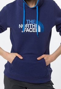 Granatowy sweatshirt z kapturem, posiadający przednią kieszeń oraz niebieskie sznurki. Zawiera duże białe logo na piersiach.