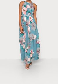 Robe maxi florale en tissu bleu, ornée de grandes fleurs roses et blanches. Encolure halter, taille cintrée et jupe fluide avec superposition transparente.