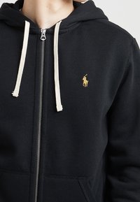 Svart zip-up hoodie i bomull med dragkedja framtill och beige snoddar. Litet gult logotyp på vänster bröstområde.