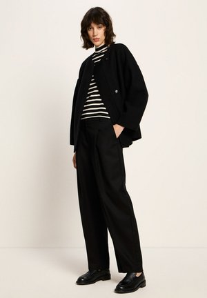 Schwarze oversized Jacke mit großen Knöpfen, über einem schwarz-weißen gestreiften Rollkragenpullover getragen, kombiniert mit hochtaillierten schwarzen Hosen und schwarzen Schuhen.