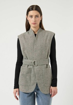 Nainen, jolla on päällään hihaton harmaa kalanruotokuvioinen liivi vyöllä, musta pitkähihainen paita, siniset farkut ja roikkuvat korvakorut, seisoo yksiväristä taustaa vasten.