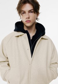Beige uldblandingjakke med afslappet pasform, trykknaplukning og sidelommer over en sort zip-up hoodie. Chinchilla-tekstur og minimalistisk design.