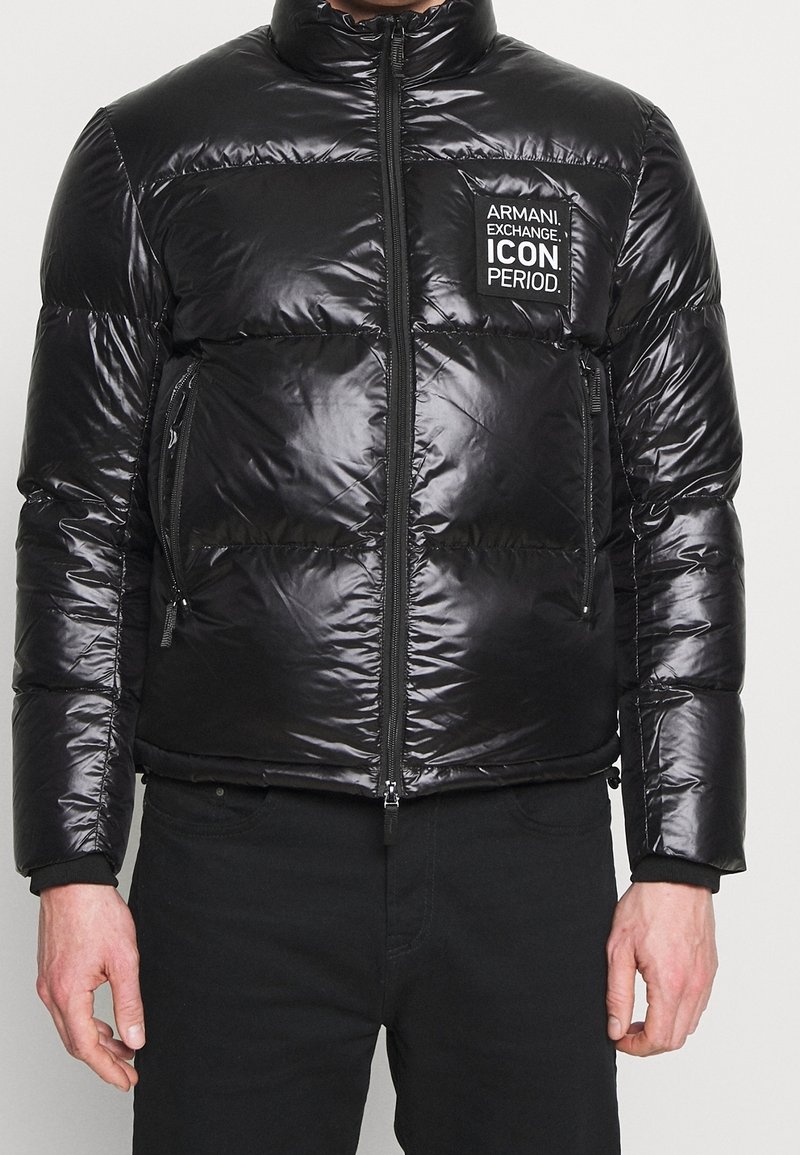 Doudoune noire brillante avec fermeture éclair devant et écusson sur la poitrine indiquant "Armani Exchange. Icon. Period.", portée par une personne debout avec un pantalon noir.