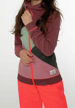 Persoon draagt een fleecejack in meerdere kleuren met roze, groen en zwarte vlakken, een oranje ritssluiting en felrode broek, en houdt de kraag van het jack vast.