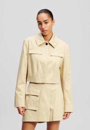 Beige cropped jas met grote voorzakken, een puntige kraag en knoopsluiting. Gladde stof met een subtiele textuur en een getailleerd ontwerp.