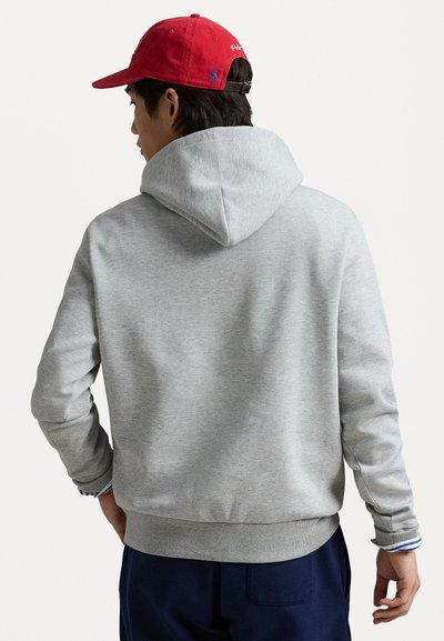 Felpa grigia con cappuccio, dal texture morbido e polsini a costine. Indossata con un cappello rosso con dettagli del logo. La vista posteriore mostra un design aderente.