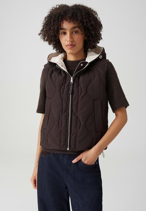 Donna con capelli ricci che indossa un gilet trapuntato scuro sopra un maglione marrone a maniche corte e jeans blu, in piedi con una mano in tasca.
