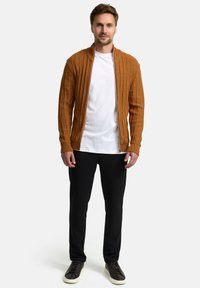 Oranger, gerippter Cardigan mit Reißverschluss über einem weißen T-Shirt, kombiniert mit schwarzer Hose und dunklen Turnschuhen. Glatte Textur, tailliertes Design und schlichte Akzente.