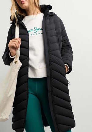 Frau trägt einen langen schwarzen gesteppter Mantel, weißen Sweatshirt, türkisfarbene Hose und trägt eine beige Tragetasche über der Schulter.