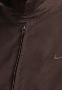 Brown Nike jacket with a high collar, featuring a zip closure, button details, and a subtle embroidered logo on the chest. Smooth texture.
