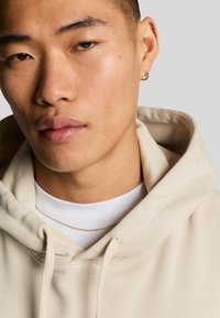 Sweat-shirt beige avec cordons, porté par-dessus un t-shirt blanc à col rond. Texture lisse, design minimaliste, avec un collier en chaîne argentée.