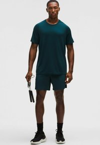 Sötét türkiz színű, rövid ujjú sportpóló, hozzá illő rövidnadrággal. A modell ugrálókötelet tart, és fekete sportcipőt visel.