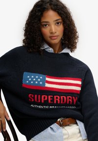Pull en tricot bleu marine avec un design texturé de drapeau américain en rouge, blanc et bleu. Le mot "SUPERDRY" est inscrit en lettres rouges en gras sous le drapeau.