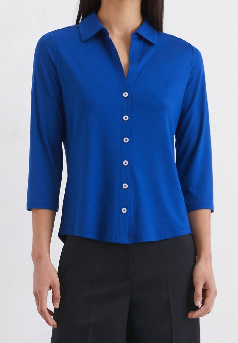 Femme portant une chemise bleue à col avec boutons, manches trois-quarts et pantalon noir sur un fond uni.