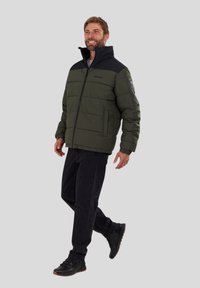 Olive green puffer jacket with a black collar, zip closure, and two side pockets; worn with black jeans and black shoes.
