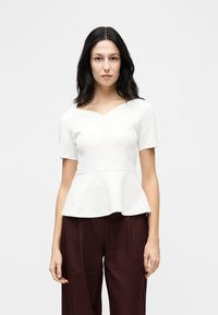 Femme aux longs cheveux noirs portant un haut péplum blanc à manches courtes et un pantalon large marron foncé, sur un fond clair uni.