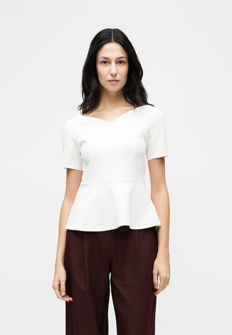 Femme aux longs cheveux noirs portant un haut péplum blanc à manches courtes et un pantalon large marron foncé, sur un fond clair uni.