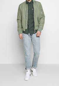 Chaqueta bomber verde oliva sobre una camisa de cuadros verde oscuro, combinada con jeans azul claro remangados y zapatillas blancas con detalles negros.