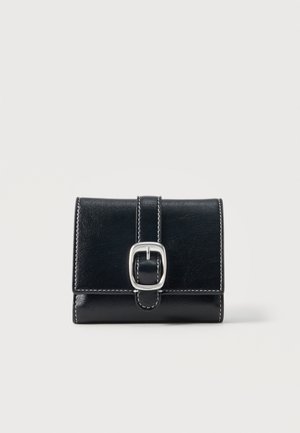 Cartera de cuero negro con costuras blancas y hebilla plateada en una correa, colocada sobre un fondo gris claro liso.